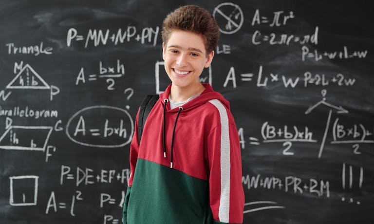 Smiling student in a classroom posing in front of a chalkboard filled with math formulas.